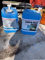 Two blue 20-litre water jugs with white nozzles placed on a concrete floor, showing some dirt and wear.