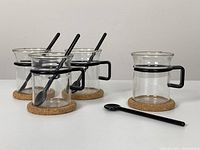 Four Bodum glass mugs with black handles, each placed on a cork coaster with a black plastic spoon inside three of the mugs and one spoon placed next to the fourth mug.