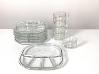 Stack of six clear square divided glass plates each with five compartments, one plate shown separately on table, alongside five stacked clear glass cups with rectangular handles and one separate cup.