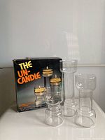 Photo showing three glass candle holders of varying heights (5, 7, 9 inches) with four floating wick holders on top, in front of original box.