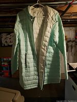 Front view of the long puffy coat hanging on a hanger in a dimly lit basement space, showing the pale green exterior quilted pattern with zipper and hood.