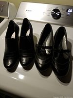 Both pairs of black women's shoes side by side on a washing machine, showing overall appearance and condition.