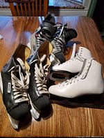 Three pairs of skates on wooden table: two black Bauer hockey skates and one pair of white Dominion Canada figure skates.