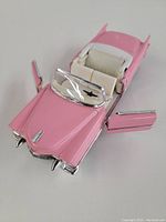 Oblique top view showing pink paint, white interior seats, open front doors, and windshield detail.