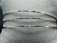 Three silver toned bangles side by side on a white cushion, showing their thin shape and etched patterns.