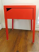 Front view of bright red Ikea Selje metal side table on wooden floor, showing the drawer closed with angled cut-out handle.