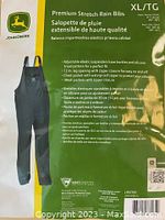 Front packaging image showing John Deere Premium Stretch Rain Bibs in black, size XL, with detailed features and product information listed.