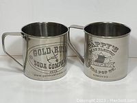 Two large metal advertising cups; one with Gold Rush Soda Company branding and the other with Pappy's Soda Pop Ribfest branding, showing front and side views.