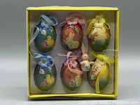 Boxed set showing six pastel-colored Easter eggs decorated with chicks and bunnies, with ribbons and flower toppers, plus a small bunny figurine in a yellow box