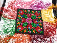Full view of the vintage Hungarian Matyó hand-embroidered square doily with multicolored silk threads on black background and colorful tatting fringe.