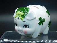 Front and side view of the white porcelain piggy bank decorated with green shamrocks and detailed painted face including lashes, smile, and pink cheeks.