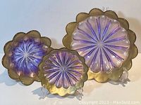 Three assorted glass bowls in lilac and gold with scalloped, fluted edges and radiating petal pattern, displayed standing up to show rim design and colors.