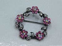 Front view of the circular brooch showing pink flower crystals with clear centers and leaf motif with clear crystals on antique finish metal base.