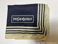 Folded view of navy and cream Yves Saint Laurent scarf showing logo inside rectangular border, hand-stitched hem visible.