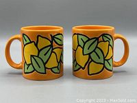 Front and side views of two orange ceramic mugs decorated with yellow lemons and green leaves on an orange background.