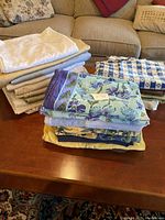 Stack of assorted tablecloths in various neutral tones including white, beige, and gray on a wooden coffee table