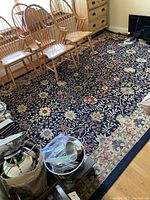 Angle view of the large black oriental style rug featuring a floral pattern, surrounded by wooden chairs and some kitchen items in the room.
