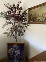 Faux ficus tree in woven basket beside framed floral print with gold frame against wall.