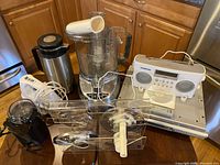 Photo showing all items in the lot: food processor, coffee thermos, coffee grinder, hand mixer, Sony CD player with remote, and Sangean radio on kitchen counter.