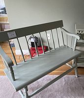 Front side angled view of the pale green wooden bench showing the backrest slats, armrest details, and turned legs.