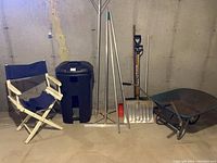Photo showing five assorted snow shovels, an extendable roof rake, an older metal wheelbarrow, a large plastic trash can on wheels, and a blue directors chair with wooden frame and fabric seat and back.