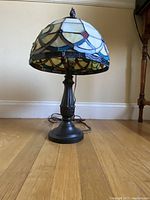 Full view of the stained glass table lamp on a hardwood floor, showing the dark metal base and stained glass shade.