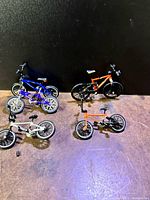 Five miniature finger bikes displayed on a surface against a dark background, some partially assembled, showing colors blue, orange, and silver.