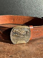 Close-up of the metal Wright Brothers belt buckle on carved leather belt, showing the embossed airplane and inscriptions.