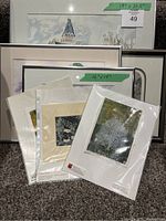 Photo of several framed artworks and unframed prints grouped together. Largest framed piece is marked 19" x 20.5", smaller frame marked 12" x 14". Four unframed prints in protective sleeves in front.