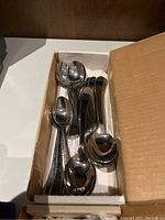 Box containing multiple stainless steel spoons, stacked inside a cardboard box with some paper lining. Spoons are standard size with a shiny finish.