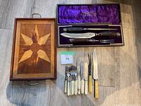 Photo of the entire lot showing the wooden tray with star inlay, velvet lined Wostenholm's carving set box with three carving tools, and an assortment of 10 knives and forks with ivory handles plus one larger knife with a wooden handle laid out on the floor.