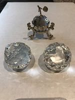 Two clear faceted Swarovski crystal ornaments beside a small hinged ornamental container with crystal top on a decorative metal stand.