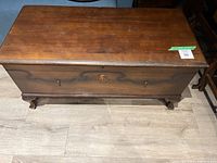 Front and top view of a vintage cedar wood trunk with carved decorative front panel and curved legs on a light hardwood floor.