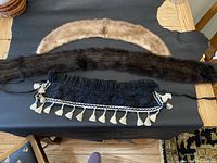 Three antique Victorian neckwear items laid flat on a black surface showing a light brown mink collar, a dark brown mink collar, and a black knitted mink neck warmer with white tassels along the edge.