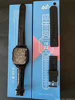 Top view of the smartwatch with attached black leather-style band and an additional black silicone replacement band next to the box insert showing watch display graphics.