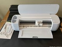 Open Cricut cutting machine with internal carriage and tool storage visible; included basic tool set on left