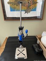 Front view of the Dremel drill press stand showing the metal base, vertical shaft, blue handle, and clamp mechanism.
