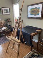 Photo showing the oak painting easel standing in a room, next to a table covered with black cloth and some framed artwork on the wall.