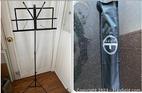 Full view of the black metal music stand fully extended in front of a white curtain, showing tripod base and adjustable arms.