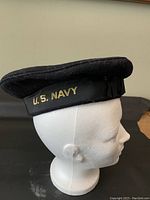Side view of dark blue/black wool U.S. Navy flat cap on mannequin head showing gold embroidered U.S. NAVY text on black band.