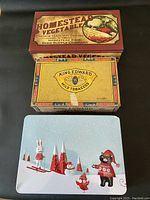 Three vintage boxes stacked: Homestead Vegetables recipe box on top, King Edward cigar box middle, winter scene cookie tin bottom.