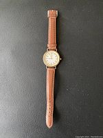 Gold-tone women's wristwatch with white dial and brown leather strap laid flat on black surface.
