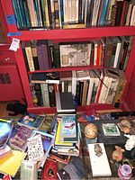 Three red bookshelves filled with various books and additional books stacked on the floor in front of the shelves.