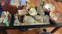 Top view of various porcelain and ceramic items on a black table, showing a repaired Chinoiserie bowl, lamp figurine, Wedgwood cup, Christmas bowl, Aynsley vase, and multiple figurines