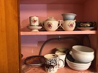 Shelving with Lenox creamer and sugar bowl, porcelain mugs with deer and floral patterns, white bowls, and decorative pieces.