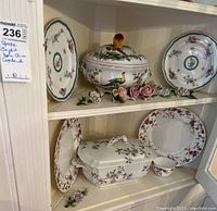 Six pieces of china displayed on shelving showing plates, serving dishes, teacup, and floral decorations.