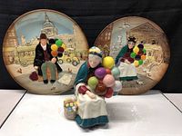 Balloon lady figurine holding colorful balloons seated in front of two decorative plates with balloon man and lady scenes.