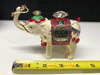 Side view of the cream-colored elephant jewel safe with colorful enamel decorations, crystal accents, and a measuring tape showing approx 5 inches length.