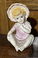 Photo of the little girl ceramic figurine showing full front view, seated position, pastel pink dress with bonnet and detailed facial features