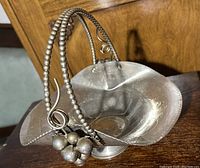 Metal candy bowl made of silver-tone metal with a decorative handle decorating with beads and metal balls. Embossed floral pattern on the bowl surface, petal-shaped edges and pedestal base.